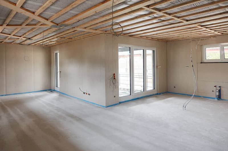 Leerer Rohbau eines Raums mit sichtbaren Deckenbalken, großen Fenstern und Kabeln an den Wänden