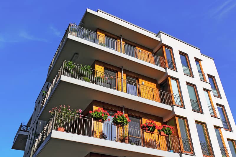 Moderner Neubau mit mehreren Balkonen, die mit Blumentöpfen und Pflanzen geschmückt sind, vor blauem Himmel