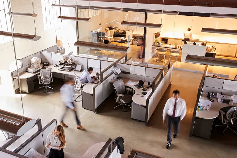 Büro mit mehreren Arbeitsplätzen, Trennwänden und Personen, die sich bewegen oder arbeiten
