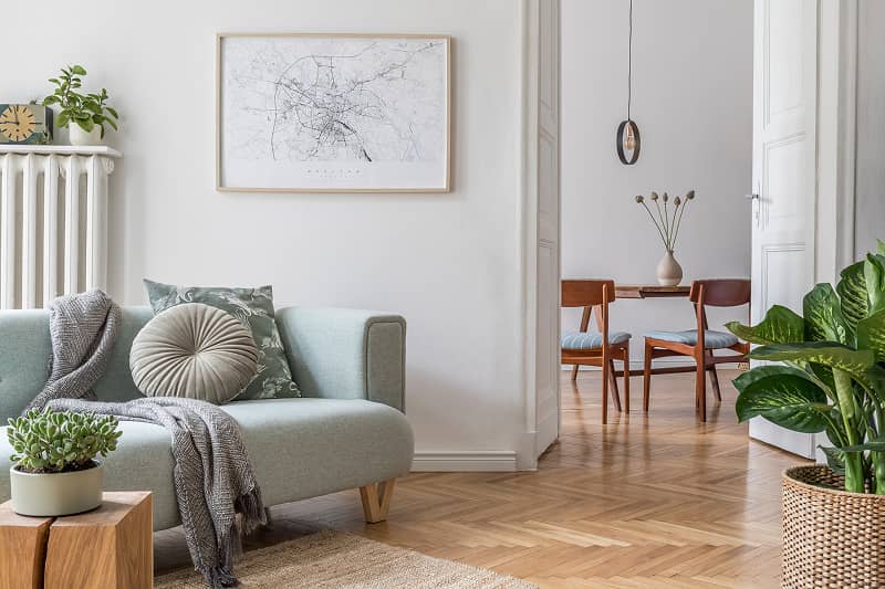 Helles Wohnzimmer mit hellgrauem Sofa, runden Kissen, Pflanzen und Holzboden, im Hintergrund Essbereich mit Holztisch und Stühlen